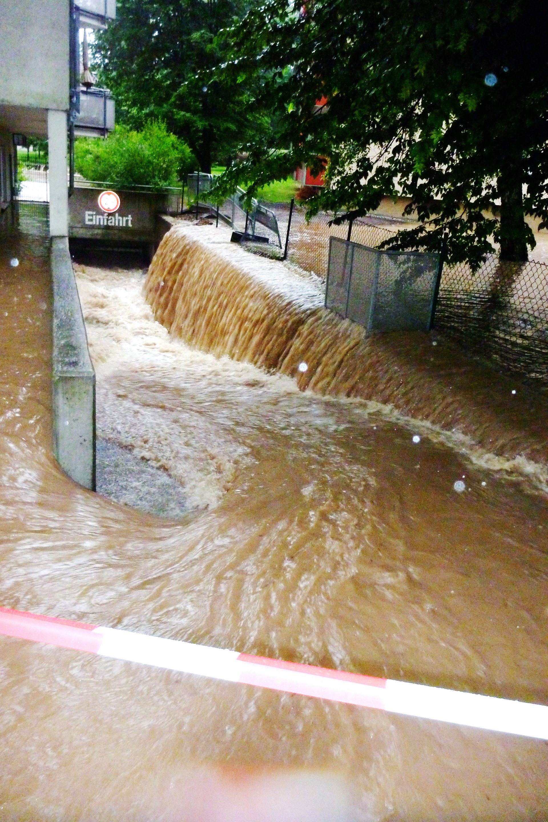 Nach einem Starkregen fließt eine Sturzflut aus schlammigem Wasser über eine Straße in den Eingang einer Tiefgarage.