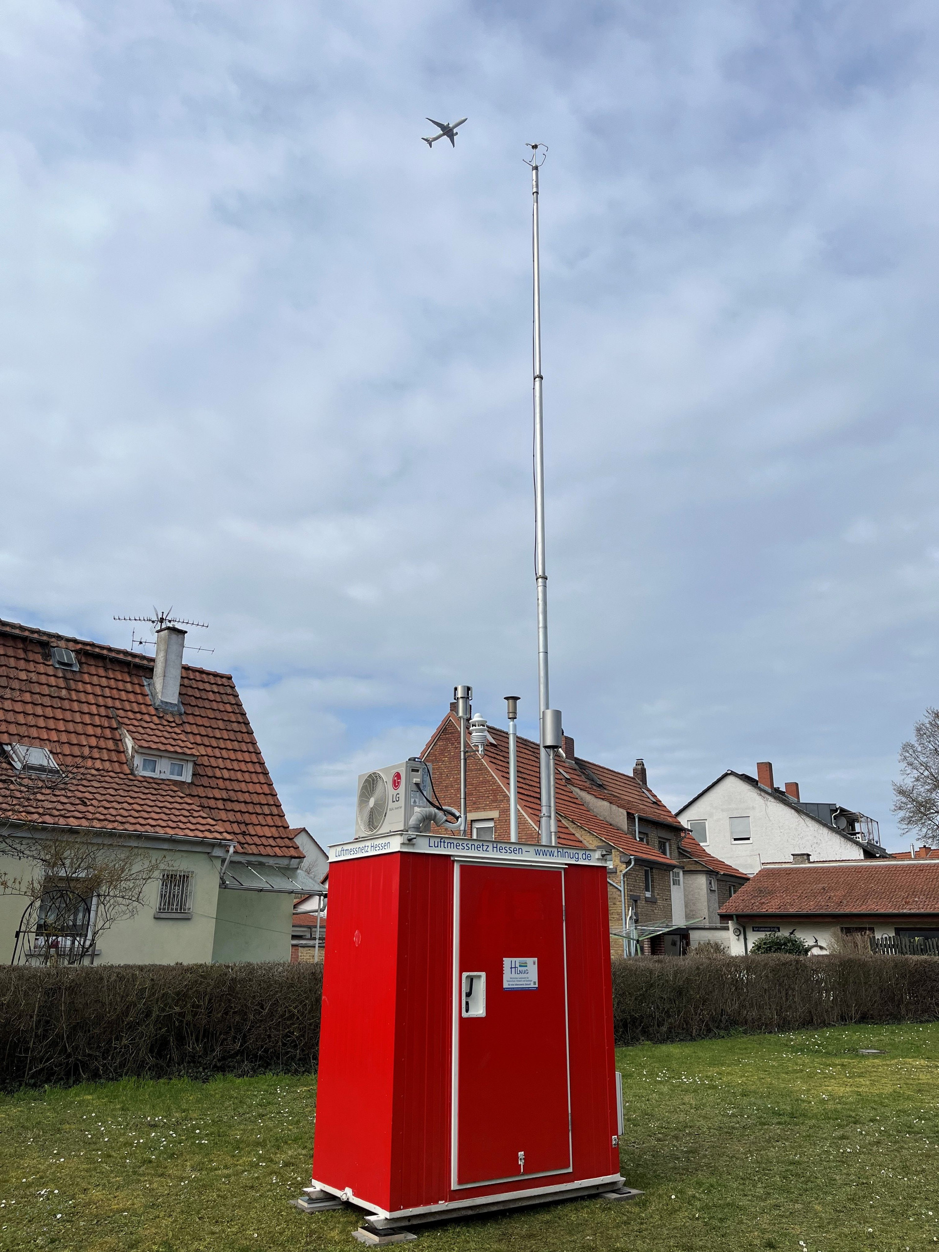 Der rote Messcontainer steht auf einer Wiese vor Wohnhäusern.