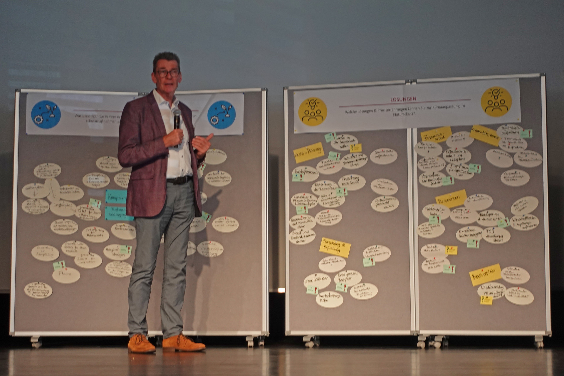 Redner steht auf der Bühne vor Pinnwänden.